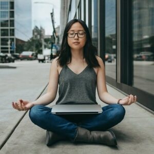 personne en pleine méditation sur un trottoir les yeux fermés et les mains ouvertes vers le ciel, un ordinateur fermé et posé sur les jambes de cette personne