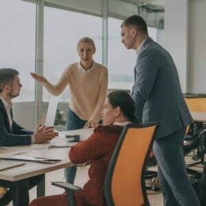 4 personnes autour d'un bureau, 2 assises, 2 debout, une des personne debout fait un signe de la main et l'autre personne debout semble mécontente, les limites entre vie professionnelle et vie personnelle se joue aussi dans la communication avec ses collégues